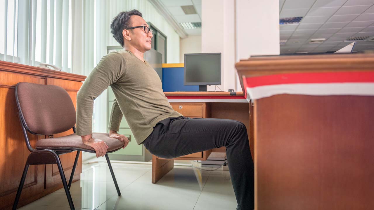 Person doing chair dips beside a desk in an office as a short exercise break.