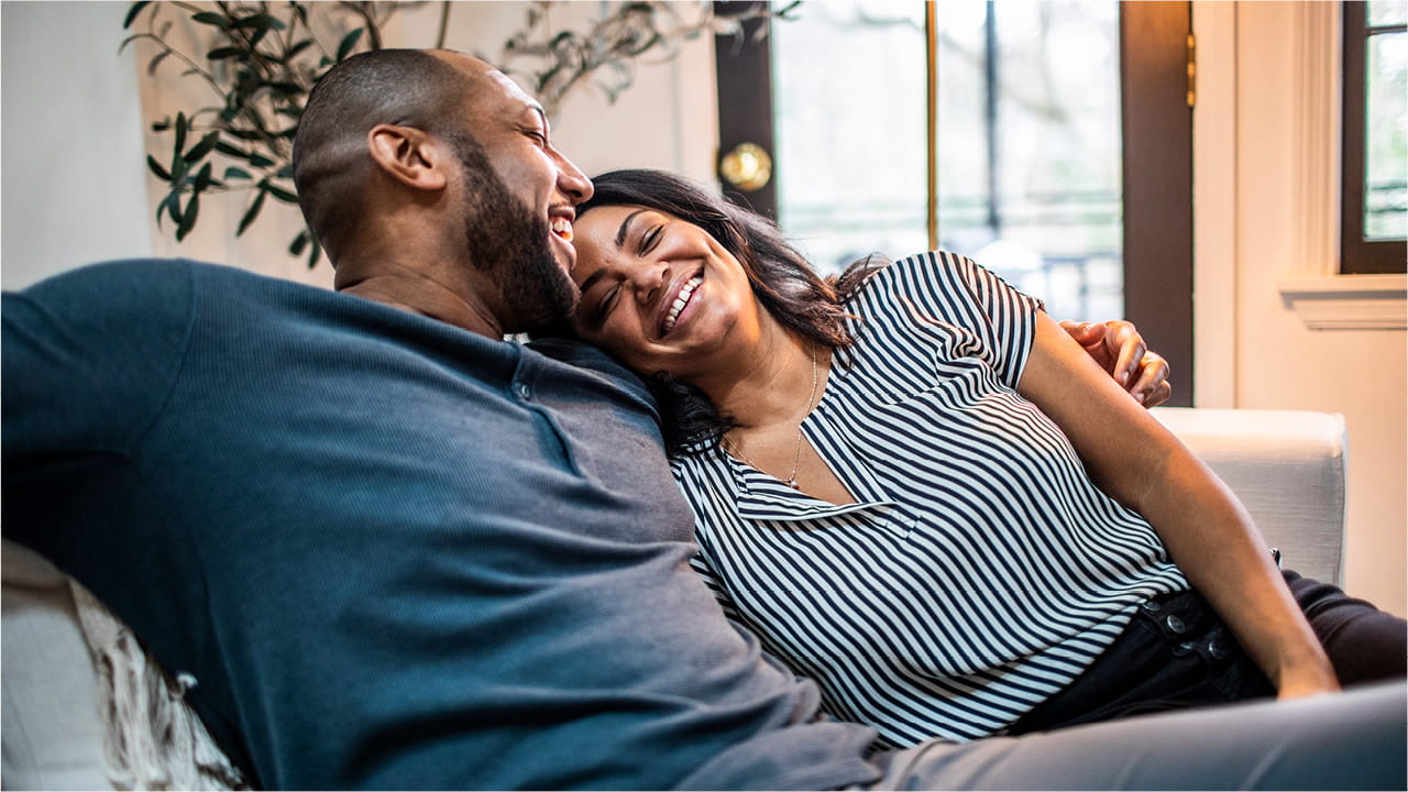 A couple sitting on a couch at home, leaning into each other and smiling in a relaxed, comfortable setting.