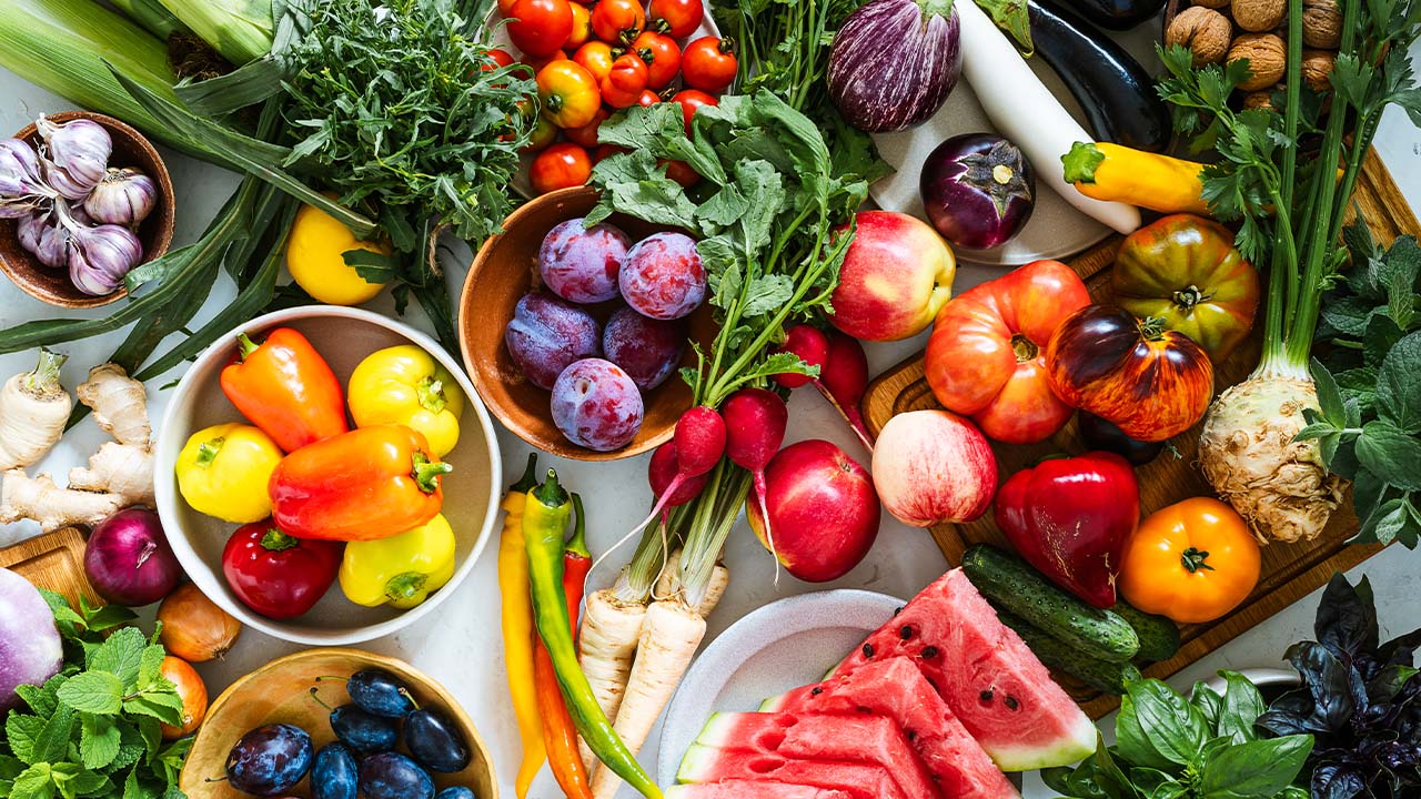 Assorted summer produce including tomatoes, peppers, radishes, watermelon, plums and fresh herbs displayed on a table