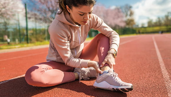 Women wearing pink athletic clothing holds her ankle in pain on a track