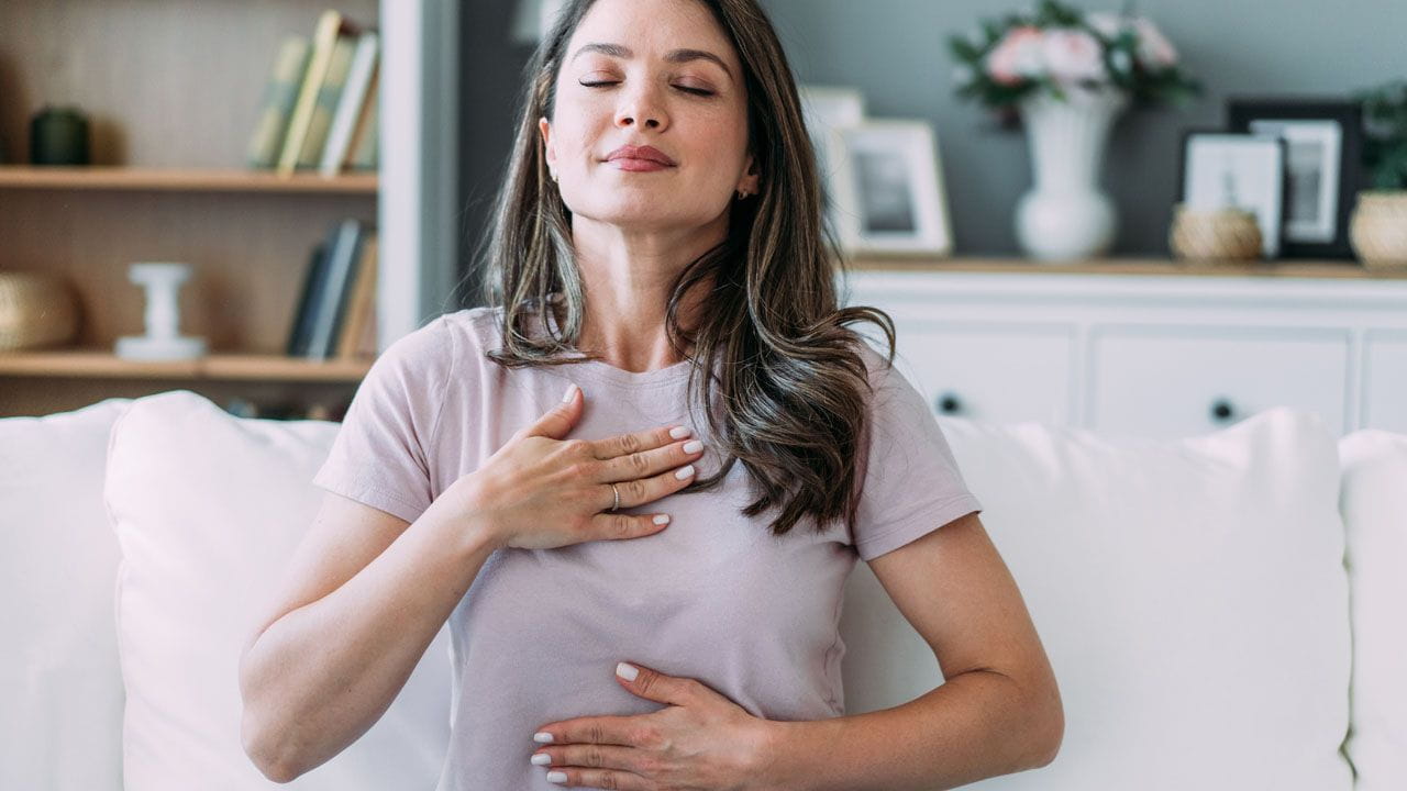Person seated indoors with one hand on the chest and one on the abdomen, demonstrating a slow breathing posture.