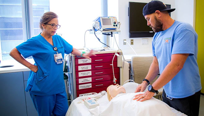 A female nurse supervises a male trainee practicing hands-on technique on a simulation mannequin.