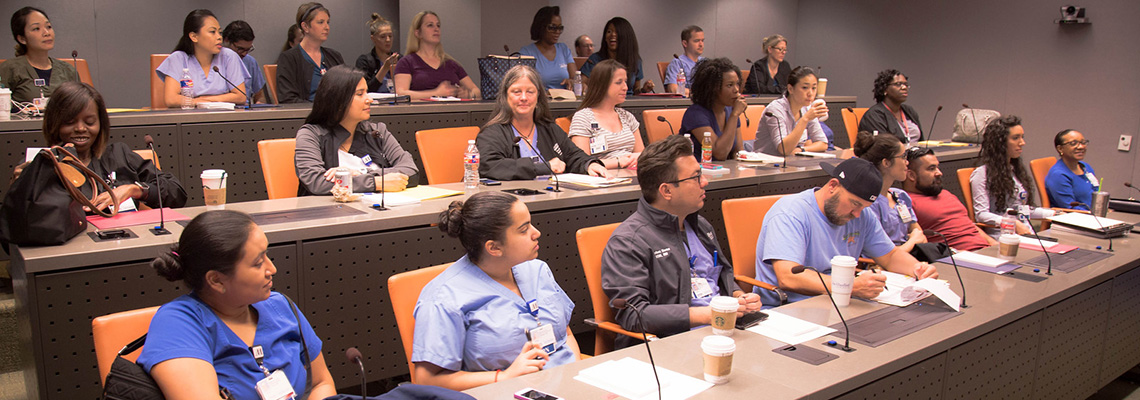 A group of men and women sit in three rows of desks in the MITIE Med Presence Suite.