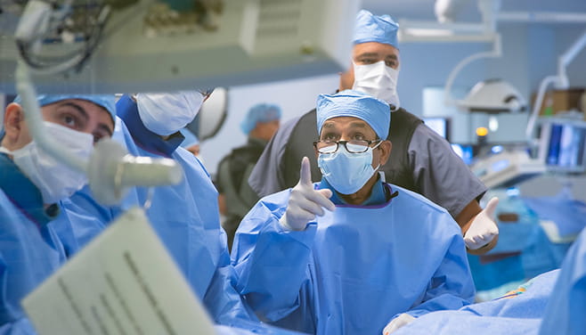 A surgeon wearing blue gown, surgical cap and mask points at a computer monitor.