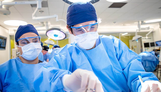 two surgeons in blue gown and scrub caps face the camera as they operate