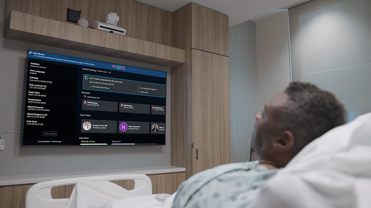 A patient lies in a bed in a hospital room reads the electronic board outlining his care during his hospital stay.