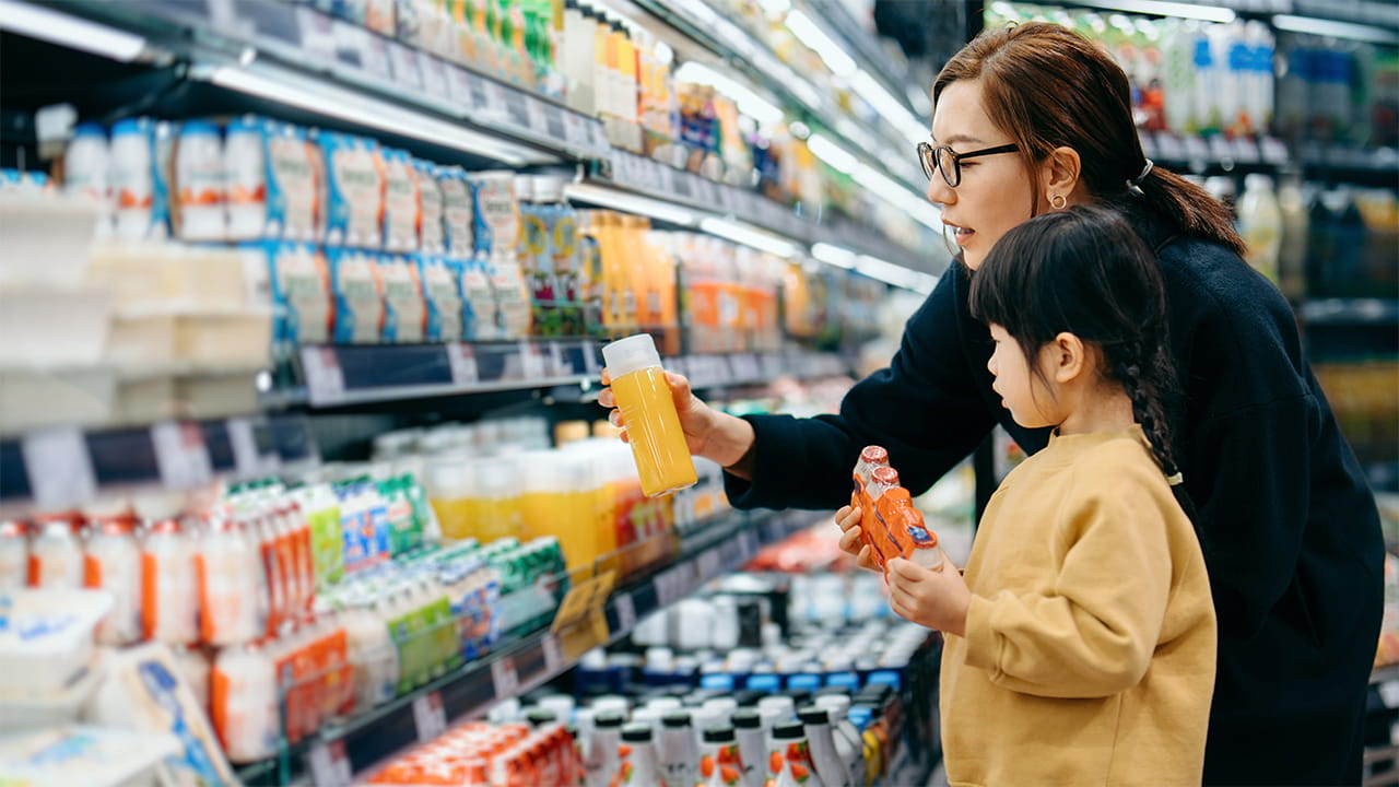 A woman and her daughter peruse the probiotic drink section of a grocery store, examining labels.