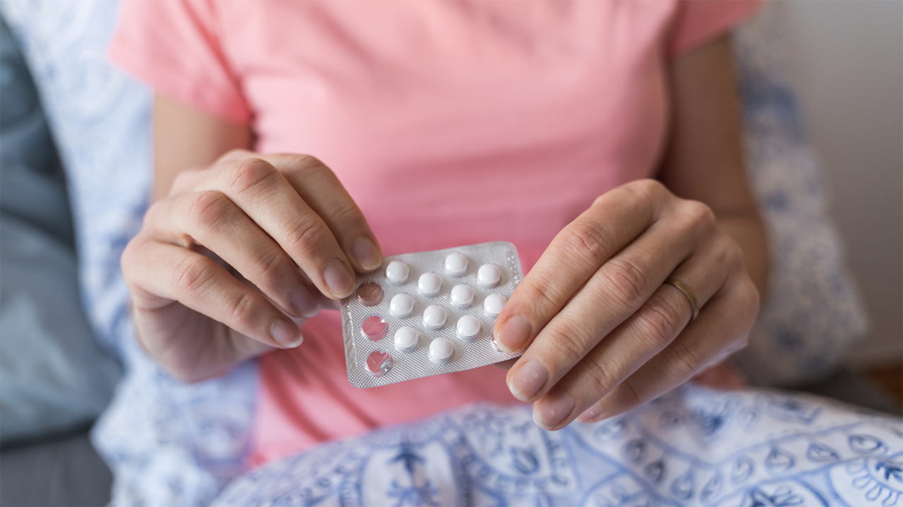 A woman in a pink shirt is lying in a bed with blue bedding holding a packet of hormone replacement therapy pills.