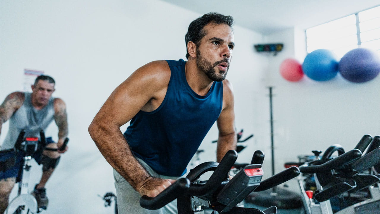A man in a blue tank exercises in a spin class with another participant in the background.