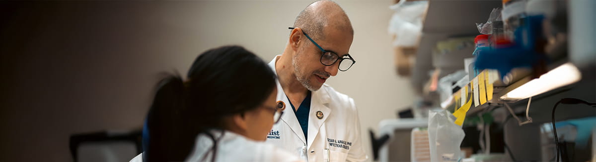 Doctor in a white lab coat and glasses reviews work at a laboratory bench beside a colleague in the foreground. Shelves with labeled supplies and research equipment line the workspace, with a shallow depth of field keeping the focus on the laboratory activity.