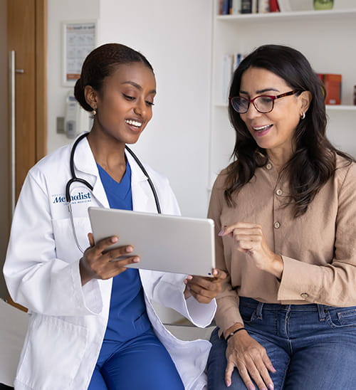 Healthcare provider wearing a white coat and stethoscope reviews information on a tablet with a patient seated beside them in a medical office.