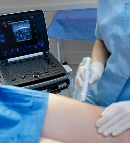 Clinician performing an ultrasound examination on a patient’s leg, with the ultrasound imaging screen visible beside the examination area.