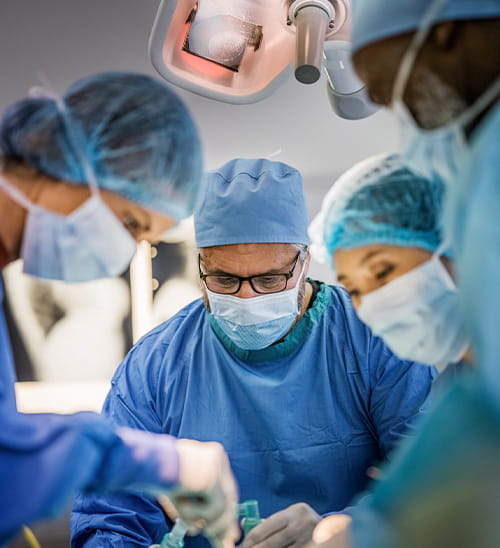 Close-up view of a surgical procedure in an operating room, showing multiple clinicians working together around a patient beneath a surgical light.