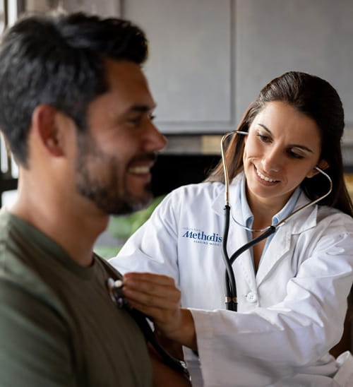 Houston Methodist healthcare provider in a white coat performing a cardiovascular examination with a stethoscope while a patient sits during an office visit.