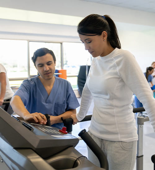 Patient walking on a treadmill during a supervised rehabilitation session, with a healthcare professional monitoring progress and adjusting equipment settings.