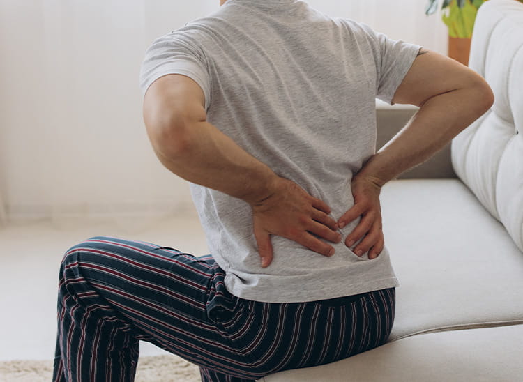 Man seen from behind sitting on the edge of a couch, holding his lower back in pain.