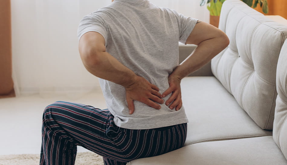 Man seen from behind sitting on the edge of a couch, holding his lower back in pain.