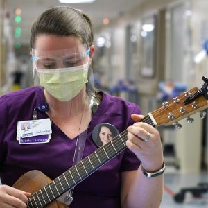 Woman in purple scrub top, mask, and face shield playing guitar on hospital unit